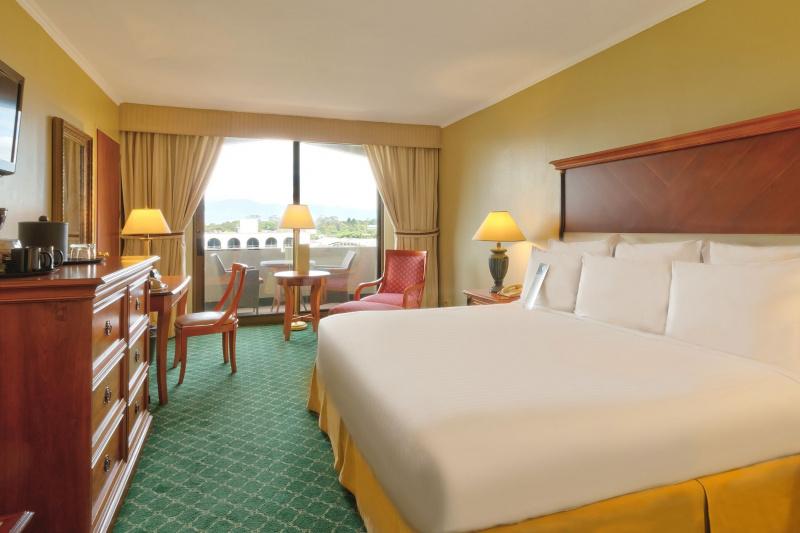 The suite has large windows, bedside tables, a desk, drawers, and an armchair.